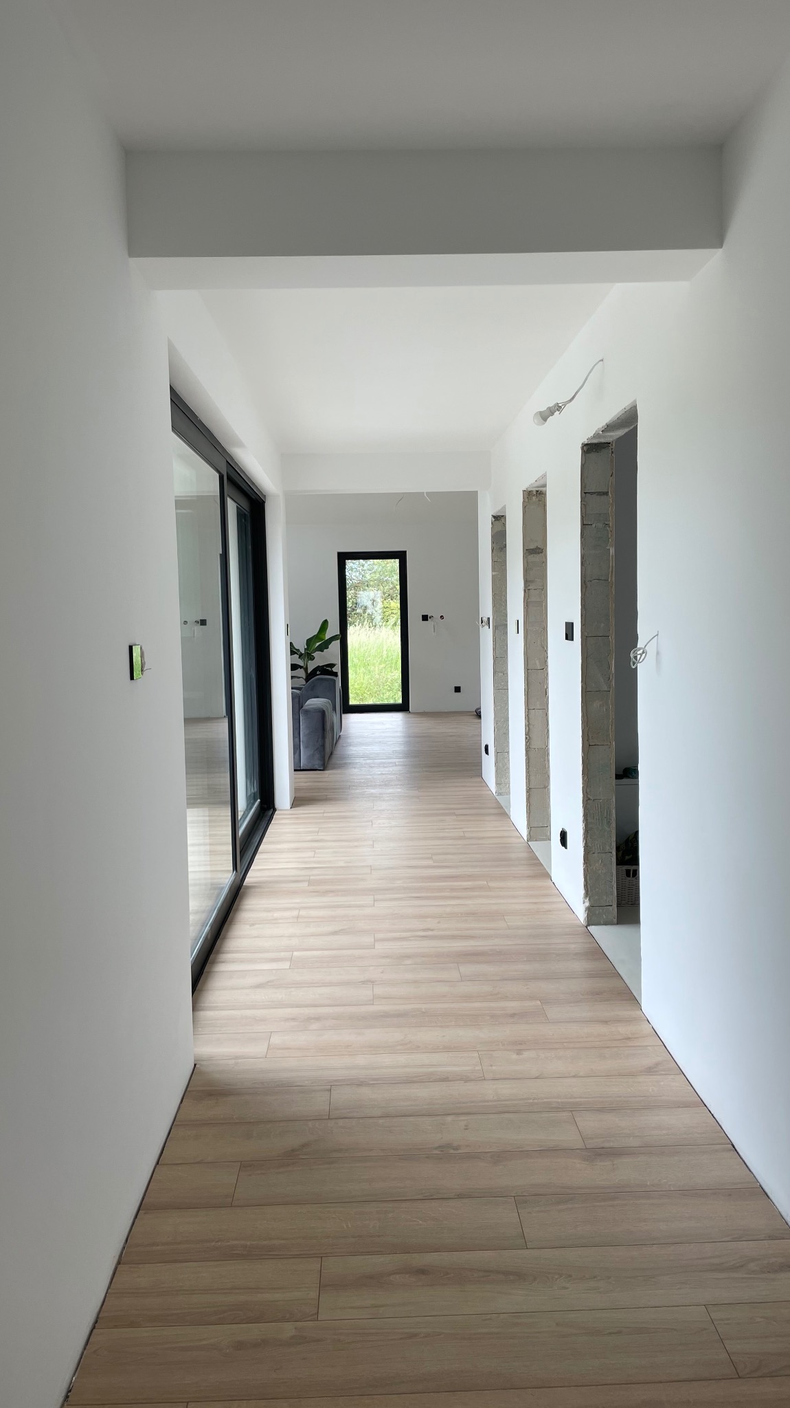 Wnętrze w trakcie wykańczania: jasne panele podłogowe imitujące drewno, surowe ściany z widocznymi otworami drzwiowymi i fragment salonu z szarą sofą w tle.