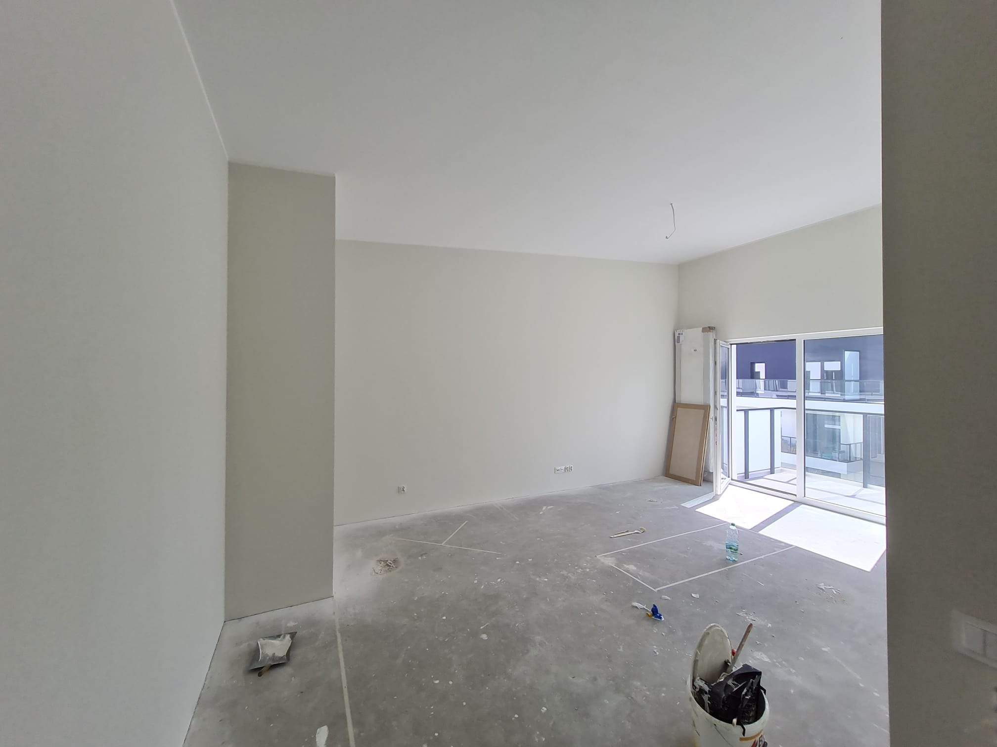 Jasne, przestronne wnętrze po szpachlowaniu ścian, z widocznymi śladami prac na podłodze i otwartym balkonem. W tle widoczne budynki.