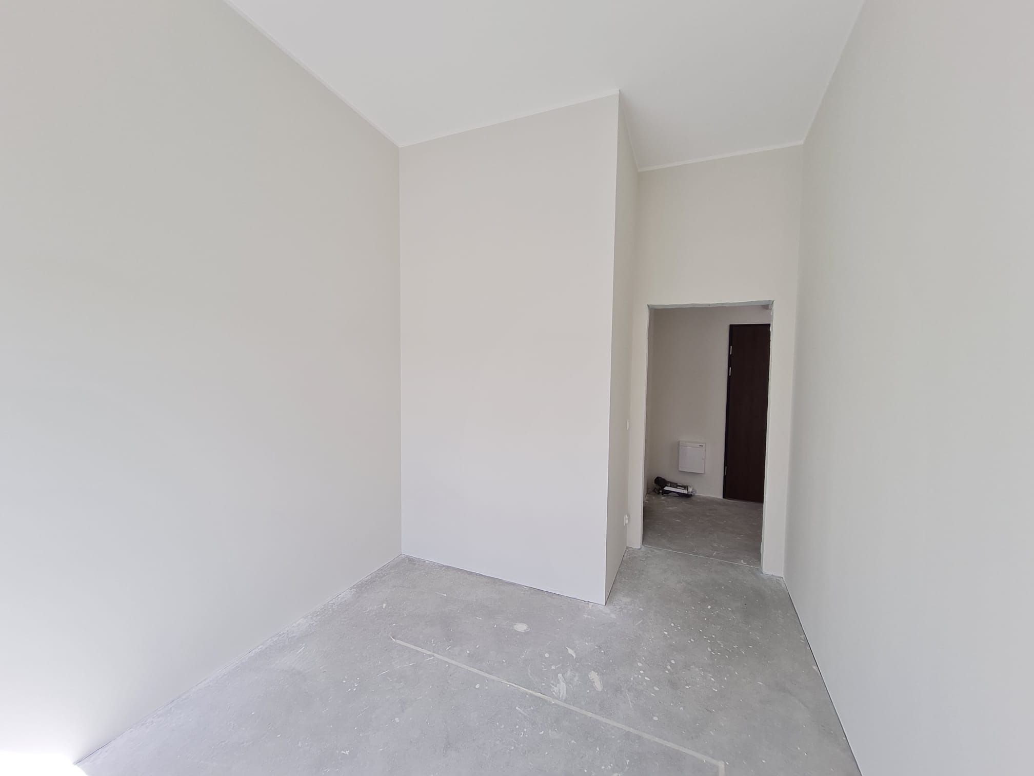 Jasne, świeżo pomalowane wnętrze pokoju z szarym betonowym podłożem i widocznym wejściem do kolejnego pomieszczenia. Minimalistyczny design, neutralna kolorystyka.