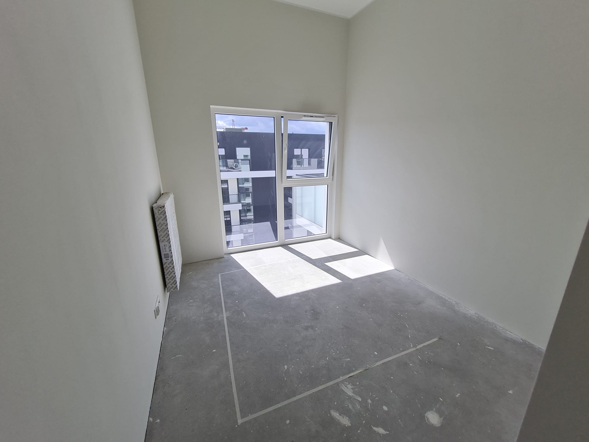 Wnętrze pokoju w stanie deweloperskim z oknem i świeżo pomalowanymi ścianami na biało. Na podłodze widoczne ślady po pracach wykończeniowych.