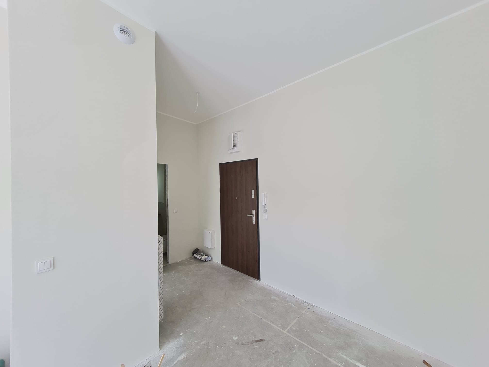 Jasne, świeżo pomalowane wnętrze z ciemnymi drzwiami i surową podłogą przygotowaną do dalszych prac wykończeniowych. Widoczny wentylator sufitowy i gniazdko elektryczne.