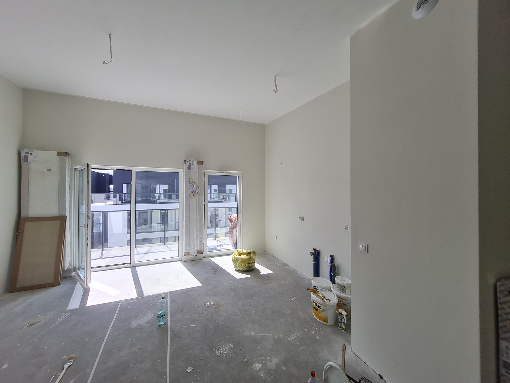 Wnętrze w trakcie wykańczania: surowa posadzka, białe ściany po gładzi, otwarte drzwi balkonowe wpuszczające światło. Widoczne narzędzia i farby malarskie.