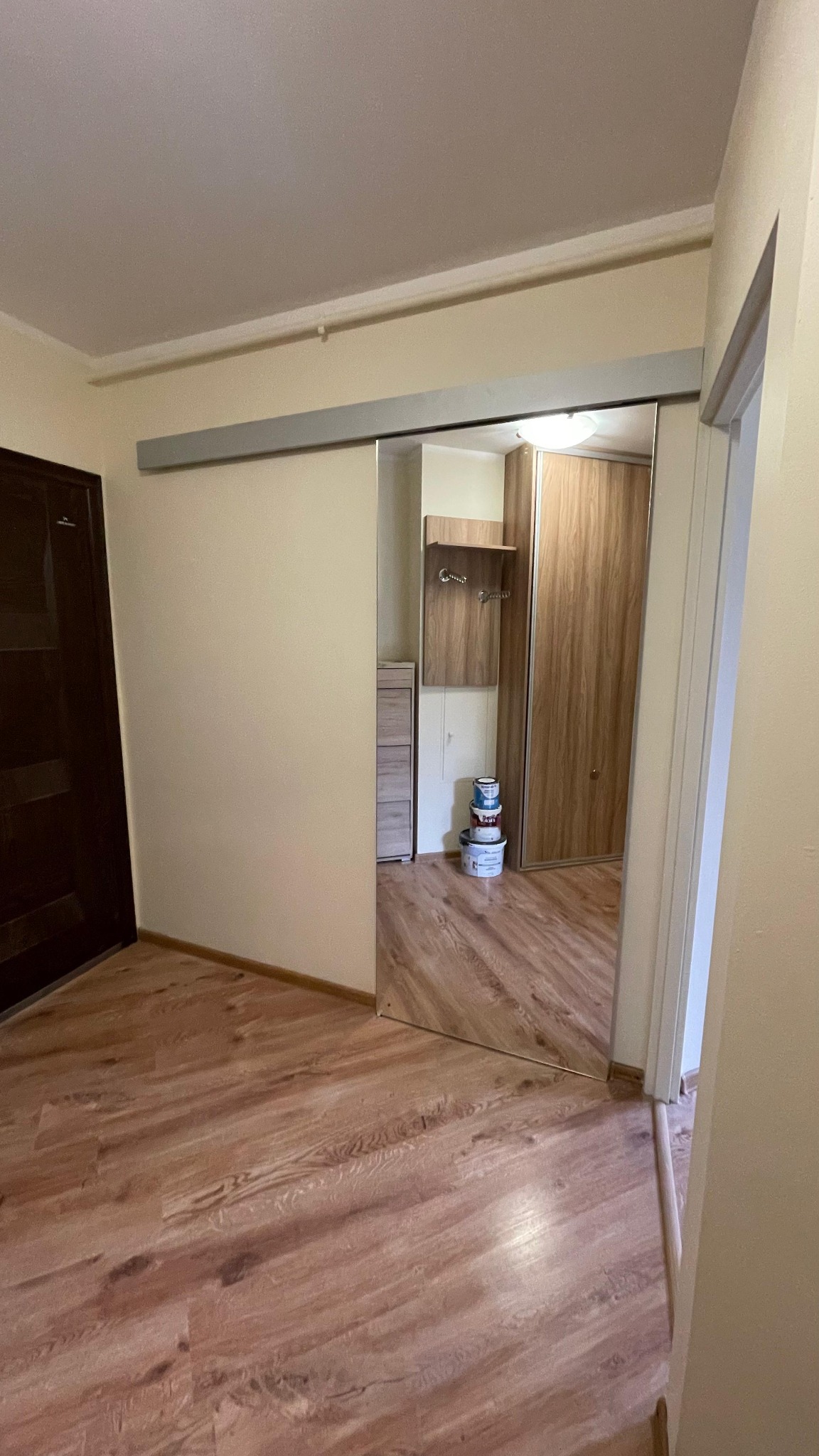 Wnętrze mieszkania z drewnianą podłogą i przesuwnymi drzwiami z lustrem, w których odbija się szafa i komoda. W rogu widoczne farby.