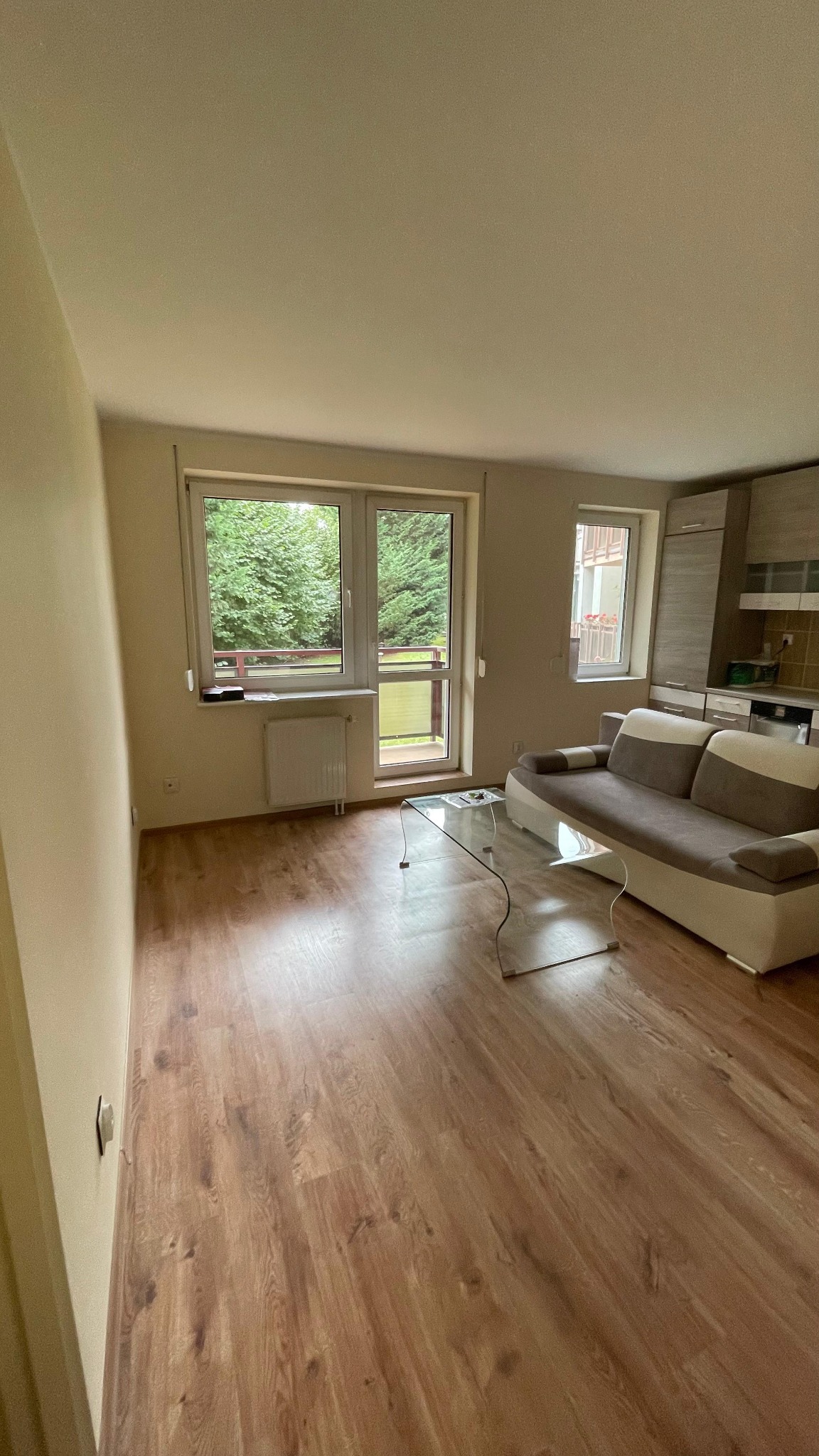 Jasny pokój z panelami podłogowymi, sofą i szklanym stolikiem. Widok na balkon i zieleń za oknem. Ukończone wnętrze po remoncie, gotowe do zamieszkania.