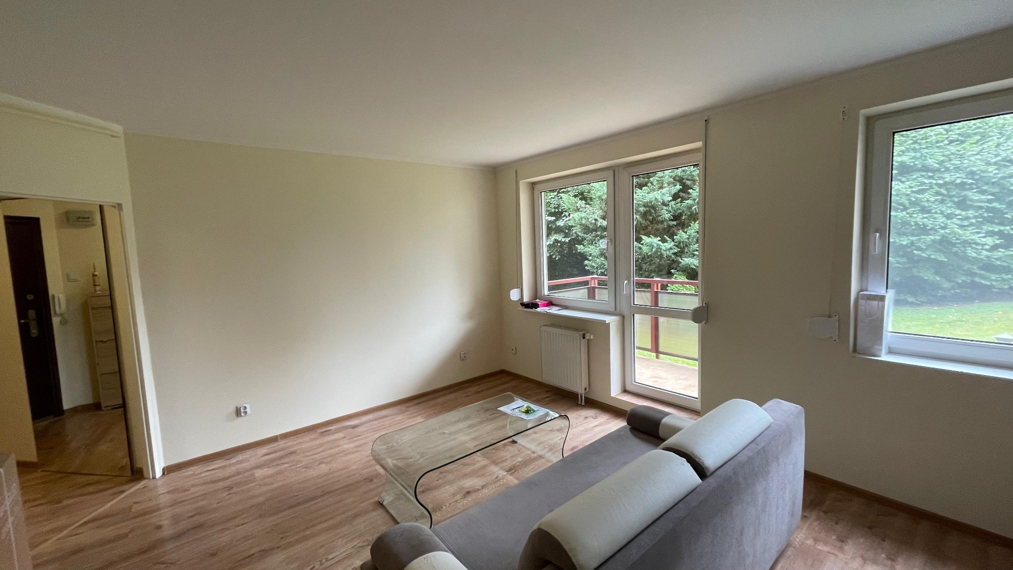 Jasne wnętrze pokoju po odświeżeniu ścian, widoczne kremowe ściany, okna z widokiem na zieleń i część sofy. Podłoga wyłożona panelami imitującymi drewno.