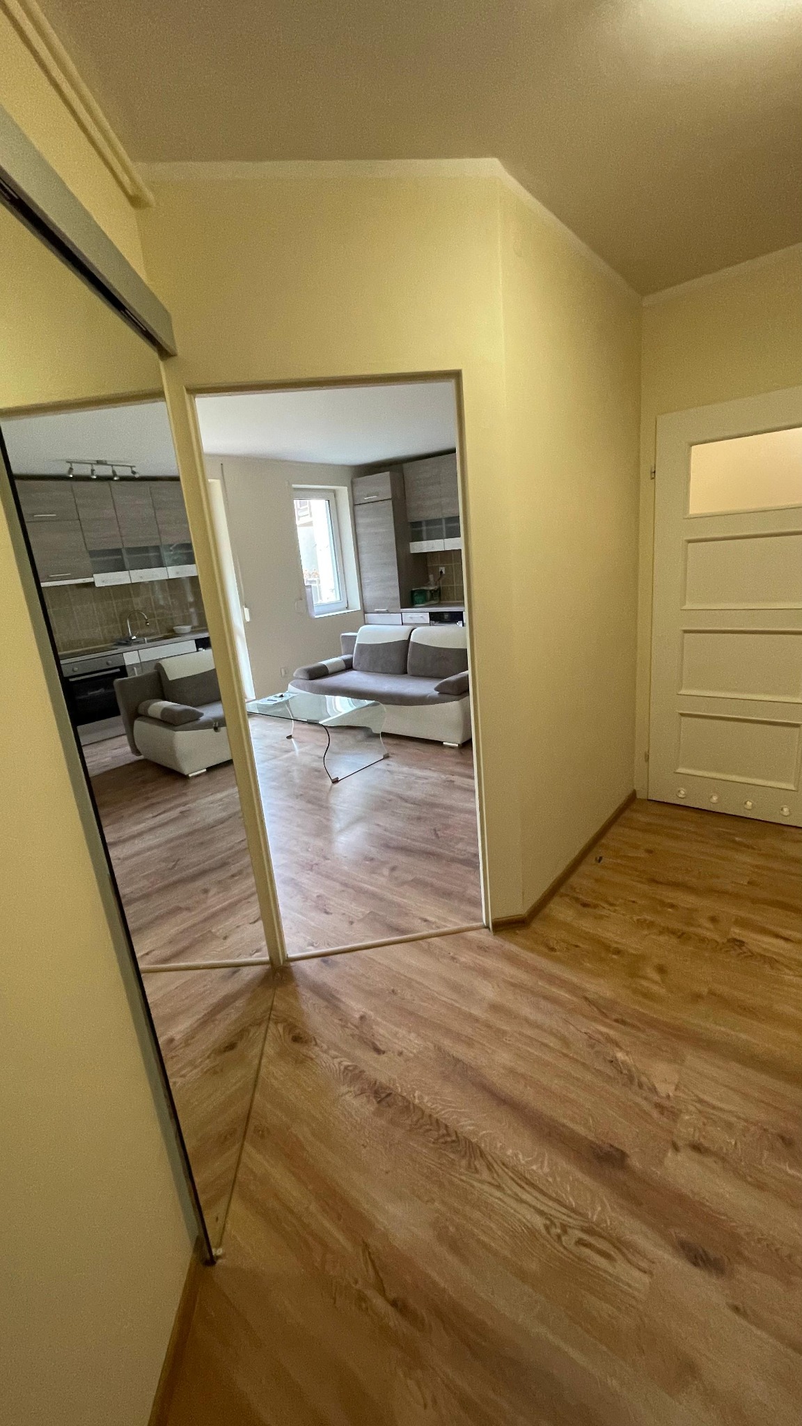 Ukończone wnętrze mieszkania: parkiet ułożony w jodełkę, kremowe ściany, widok na salon z szarą sofą i kuchnię w tle. Lustro w szafie odbija przestrzeń.