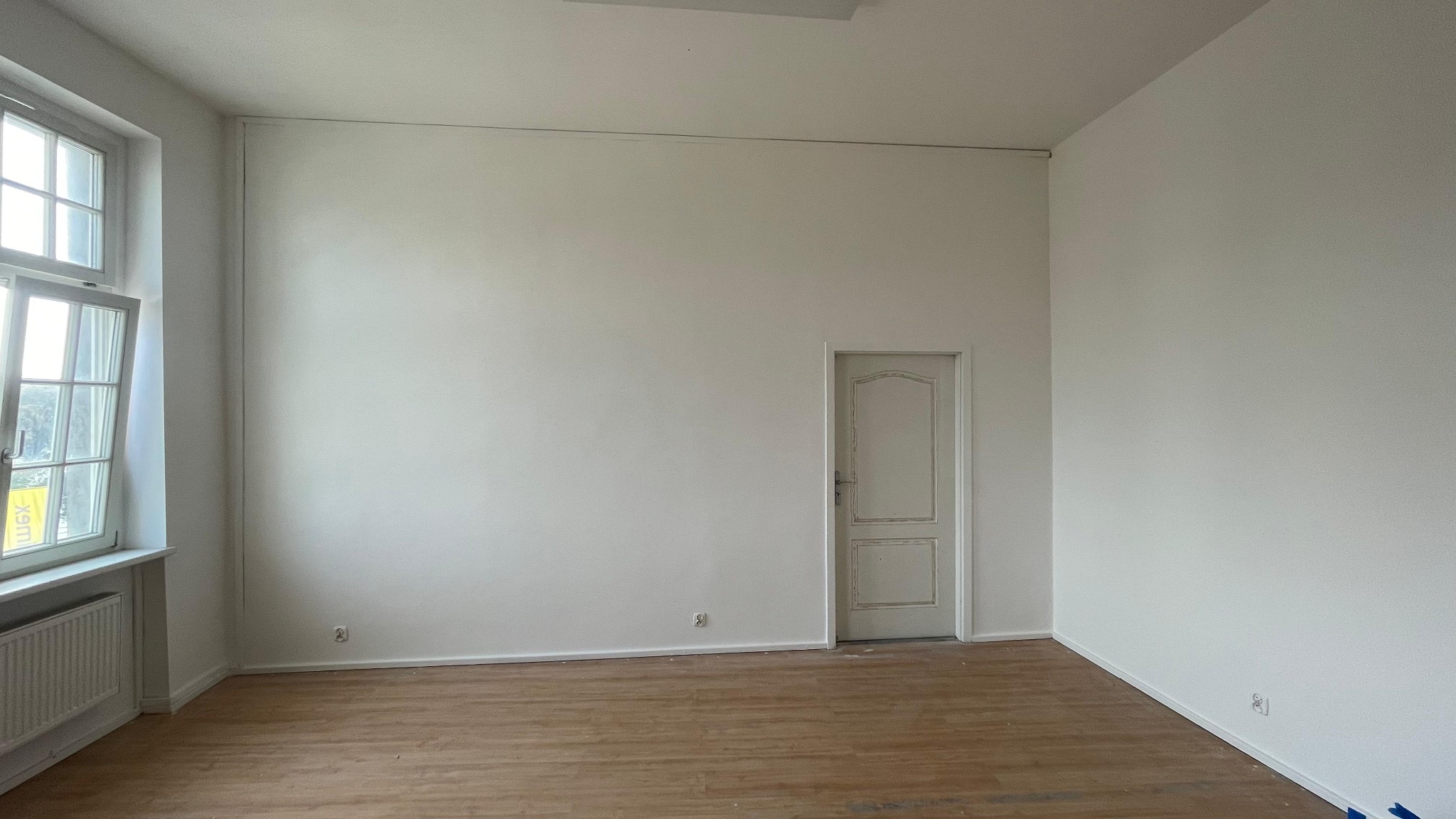 Jasne, odświeżone wnętrze studia fotograficznego z drewnianą podłogą, białymi ścianami i oknem po lewej stronie. W tle widoczne drzwi.