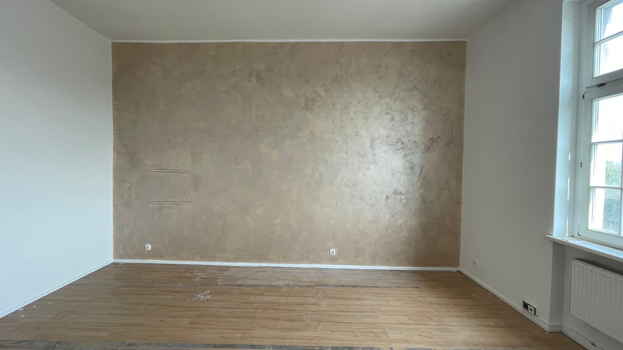 Pokój po malowaniu: ściana z efektem dekoracyjnym w odcieniach beżu, białe ściany boczne, drewnopodobna podłoga, okno z grzejnikiem. Widoczne gniazdka elektryczne.