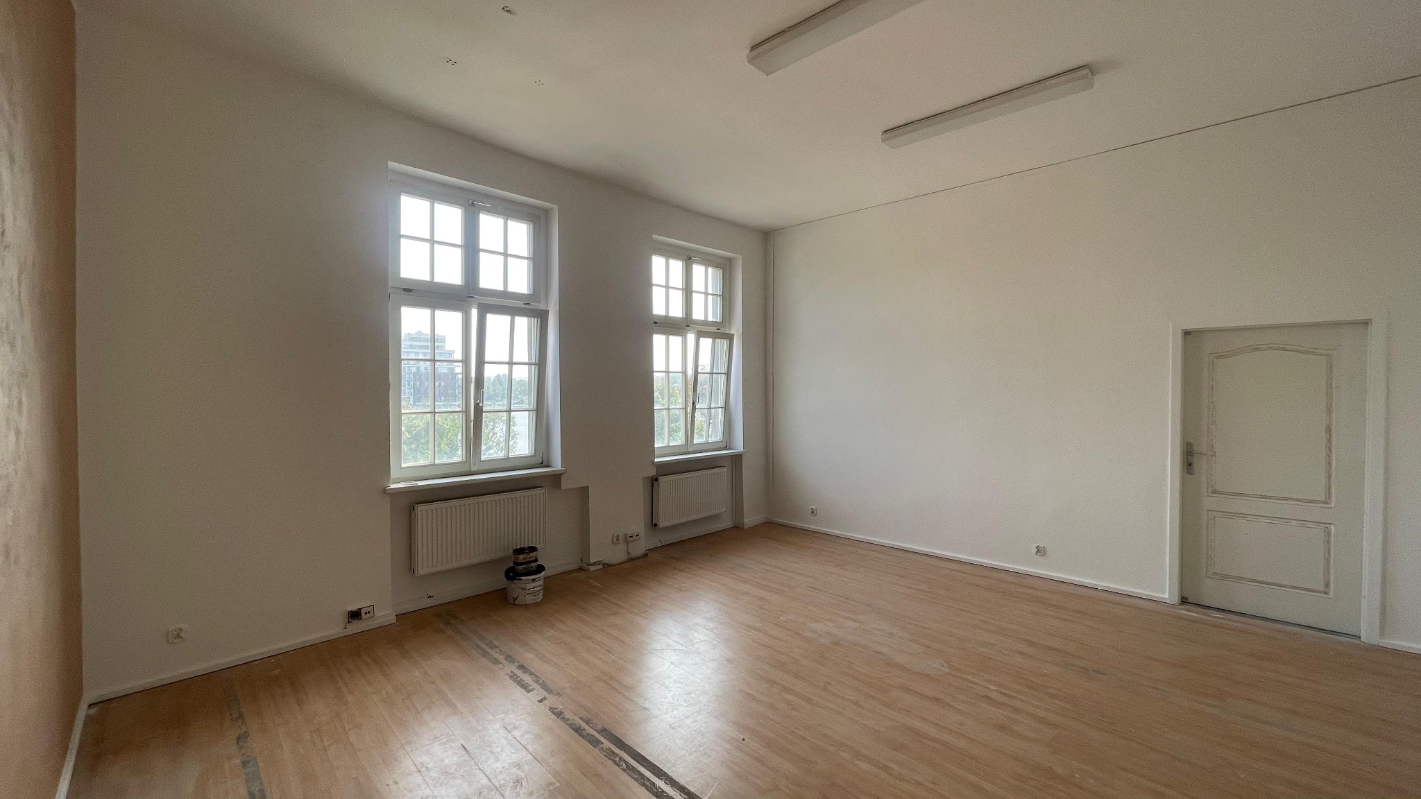 Jasne, puste pomieszczenie z dwoma oknami, drewnianą podłogą i świeżo pomalowanymi ścianami. Na podłodze widoczne puszki z farbą, sugerujące etap prac wykończeniowych.