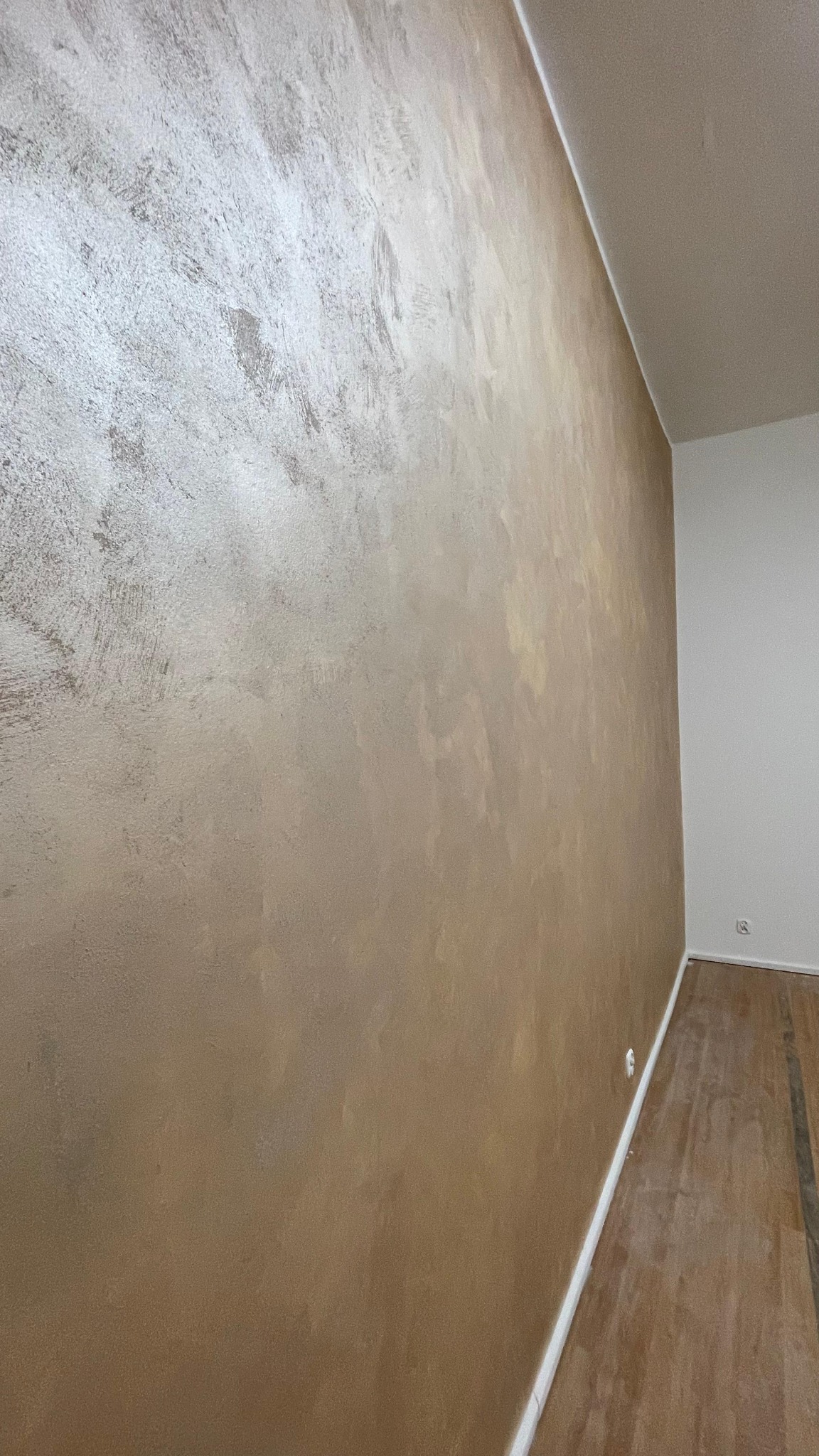 Ściana z efektem dekoracyjnym w odcieniach beżu i srebra, widoczna pod kątem w jasnym wnętrzu. Na podłodze parkiet, a w rogu biała ściana.