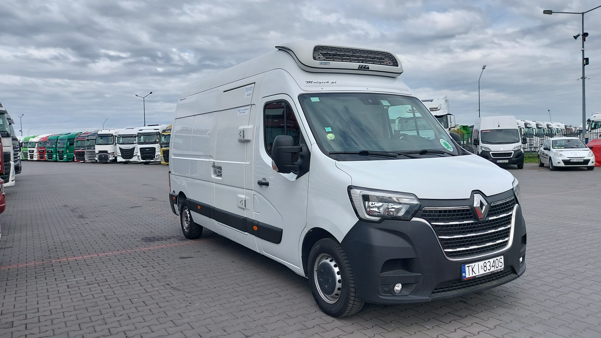 Biały, chłodniczy Renault Master na parkingu z rzędami ciężarówek w tle, pochmurne niebo. Widoczna tablica rejestracyjna i logo firmy transportowej na pojeździe.