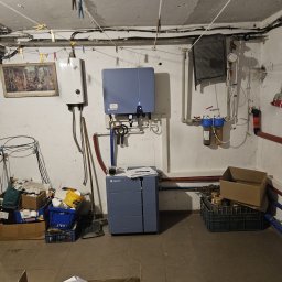 IQDOM24 Sp. z o.o. - Instalacja Solplanet w piwnicy: szara obudowa, rury, filtry wody. Na górze wisi suszarka, a na ścianie obraz. W tle widoczne kartony i przedmioty.