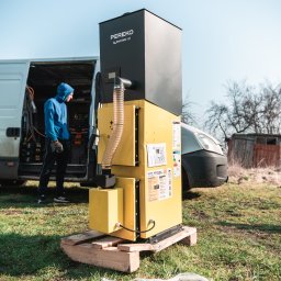 IQDOM24 Sp. z o.o. - Żółto-czarny piec Pereko QmPell EVO Bio na palecie, z pracownikiem w tle przy vanie. Widoczne ulotki i naklejki informacyjne na piecu. Instalacja na trawie.