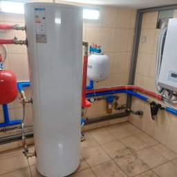 IQDOM24 Sp. z o.o. - Biały zasobnik ciepłej wody użytkowej z podłączonymi rurami miedzianymi i plastikowymi w kotłowni, obok biała pompa ciepła Kaisai na ścianie z beżowych płytek.