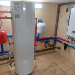 Biały zasobnik ciepłej wody użytkowej z podłączonymi rurami miedzianymi i plastikowymi w kotłowni, obok biała pompa ciepła Kaisai na ścianie z beżowych płytek.