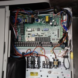 IQDOM24 Sp. z o.o. - Otwarta obudowa pompy ciepła z widoczną elektroniką, licznymi przewodami i złączami. Widoczna tabliczka ostrzegawcza i fragment pompy.