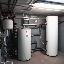 IQDOM24 Sp. z o.o. - Dwa białe zbiorniki akumulacyjne w szarej kotłowni, połączone rurami z izolacją. Widoczne liczne przewody pod sufitem i skrzynka elektryczna na ścianie.