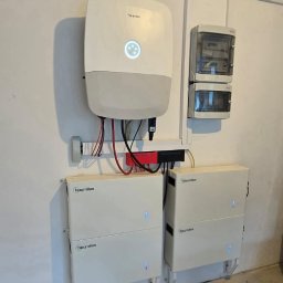 IQDOM24 Sp. z o.o. - Biały inwerter Hoymiles z podświetlanym wskaźnikiem i dwa moduły akumulatorów Hoymiles, zamontowane na białej ścianie obok skrzynek elektrycznych. Widoczne okablowanie.