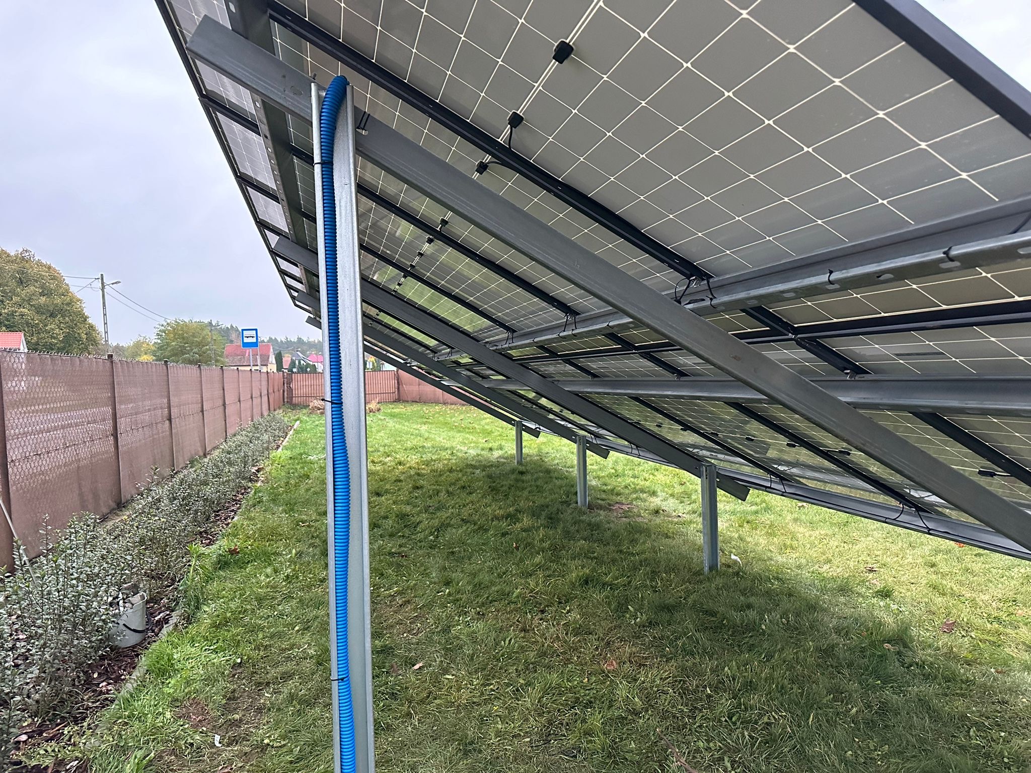 Montaż paneli fotowoltaicznych na gruncie, widok od spodu. Konstrukcja wsporcza z metalu, niebieska rura osłonowa kabli. Trawa i ogrodzenie w tle.