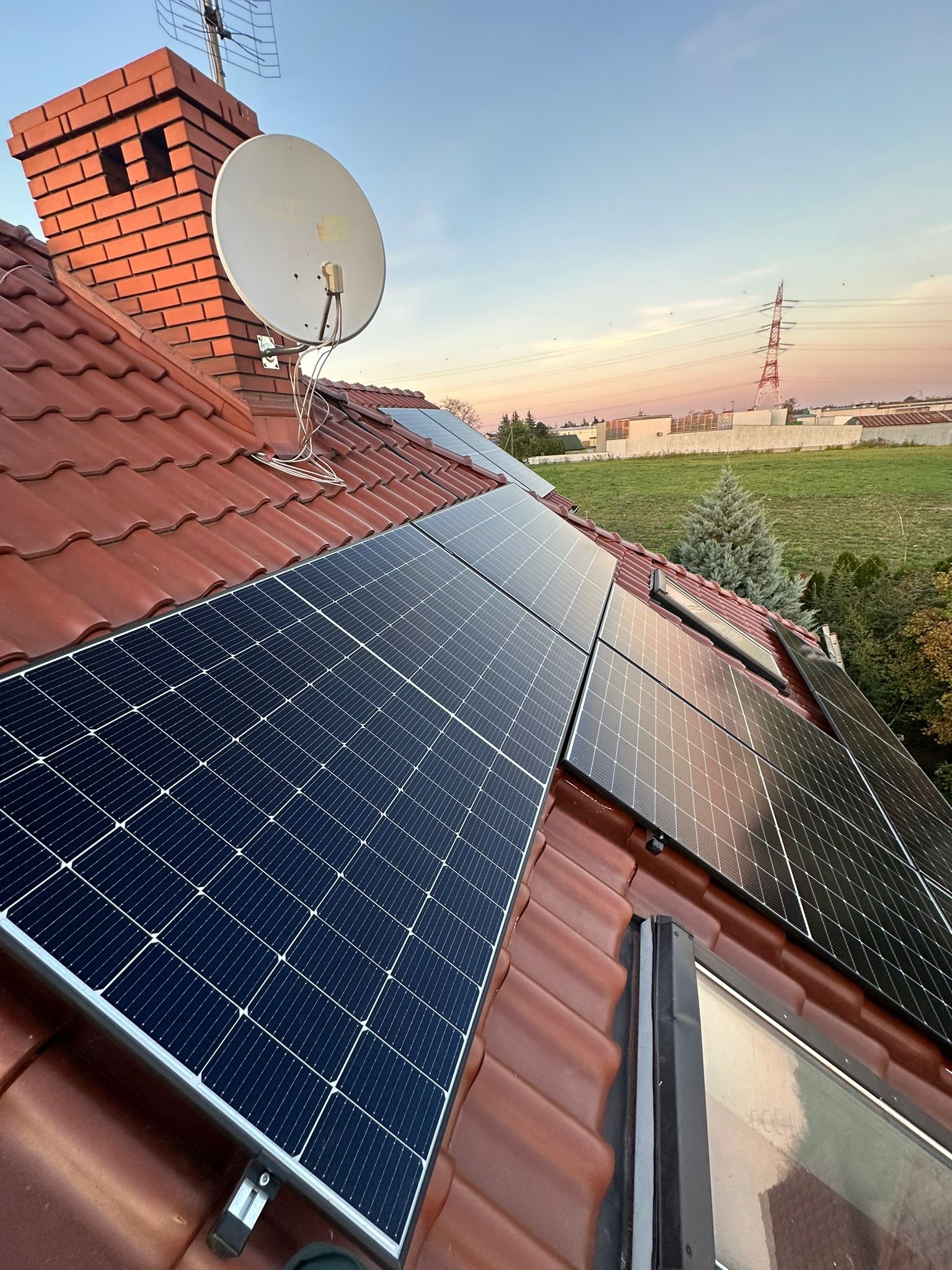 Instalacja fotowoltaiczna na dachu z czerwoną dachówką, kominem i anteną satelitarną. W tle słup wysokiego napięcia i zachodzące słońce.