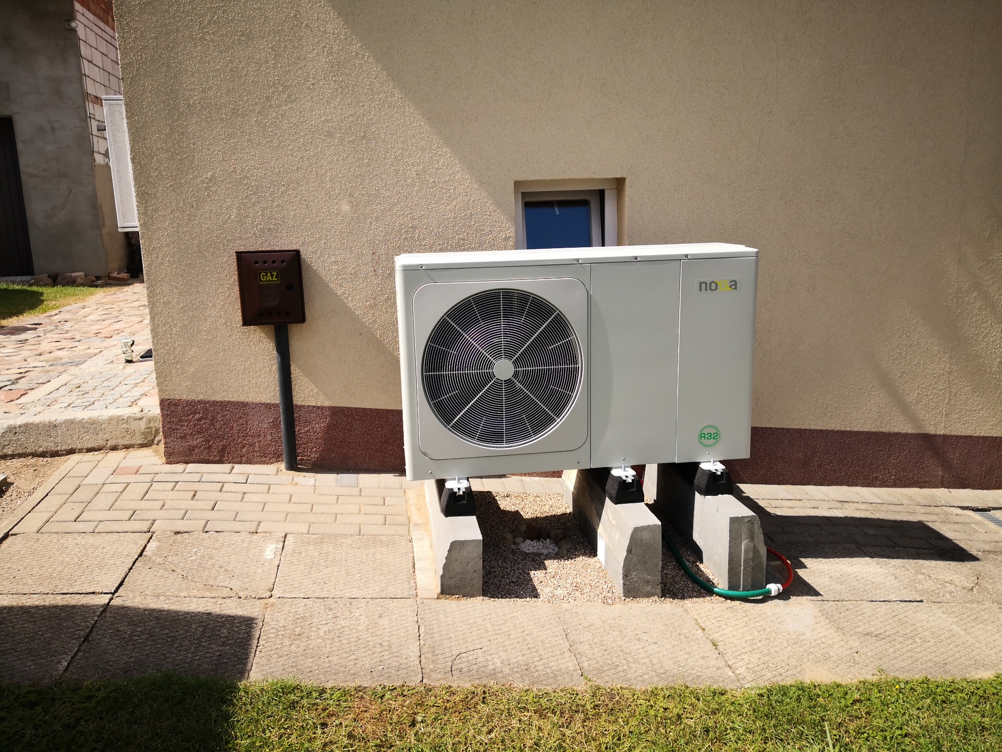 Zewnętrzna jednostka pompy ciepła Nova, zamontowana na betonowych blokach przy beżowej ścianie budynku z brązowym cokołem i skrzynką gazową.