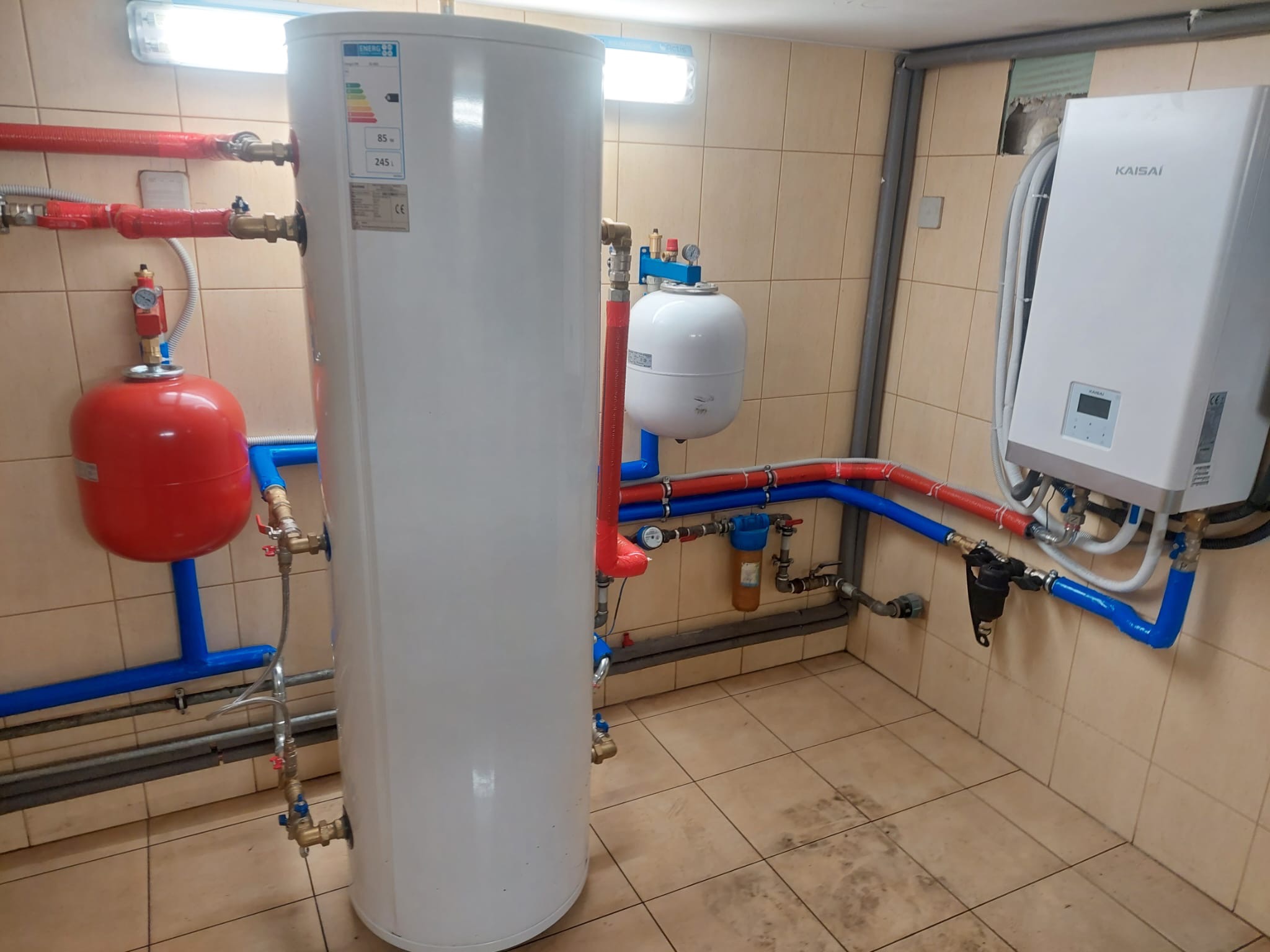 Biały zasobnik ciepłej wody użytkowej z podłączonymi rurami miedzianymi i plastikowymi w kotłowni, obok biała pompa ciepła Kaisai na ścianie z beżowych płytek.