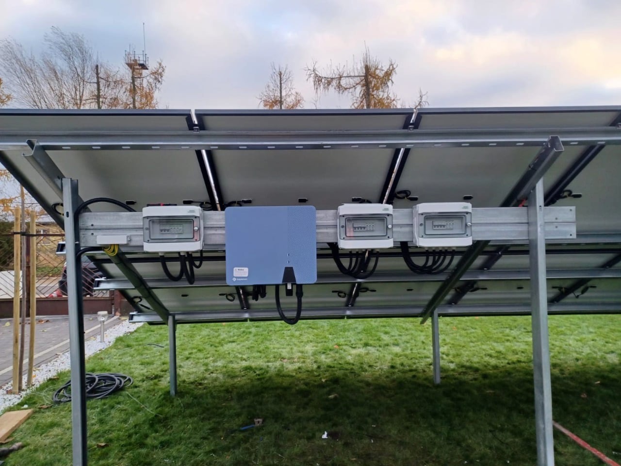 Montaż paneli fotowoltaicznych od spodu, widoczne kable, skrzynki elektryczne i konstrukcja wsporcza na tle zielonego trawnika i drzew z jesiennymi liśćmi.