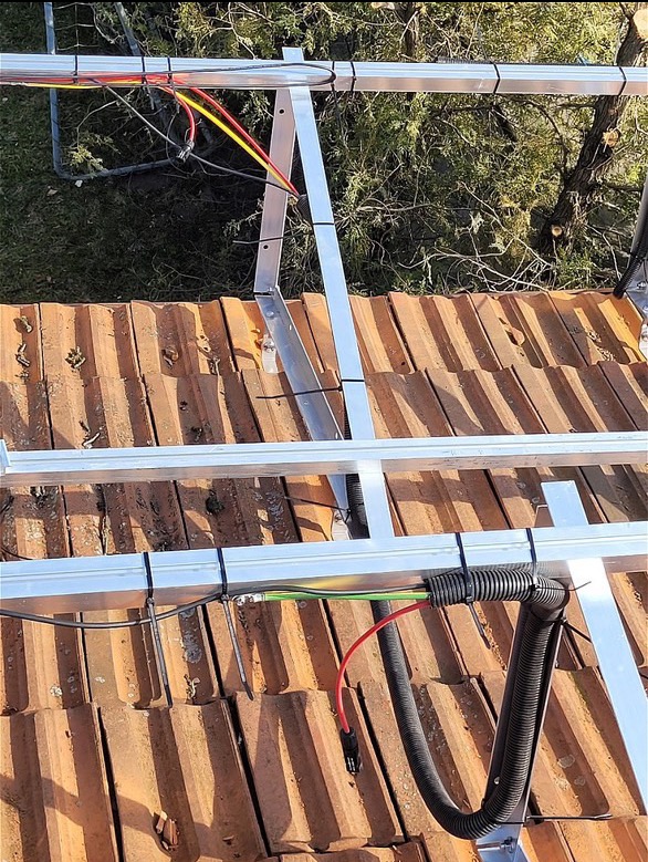 Montaż przewodów instalacji fotowoltaicznej na dachu pokrytym czerwoną dachówką. Widoczne aluminiowe profile i peszel z kablami.