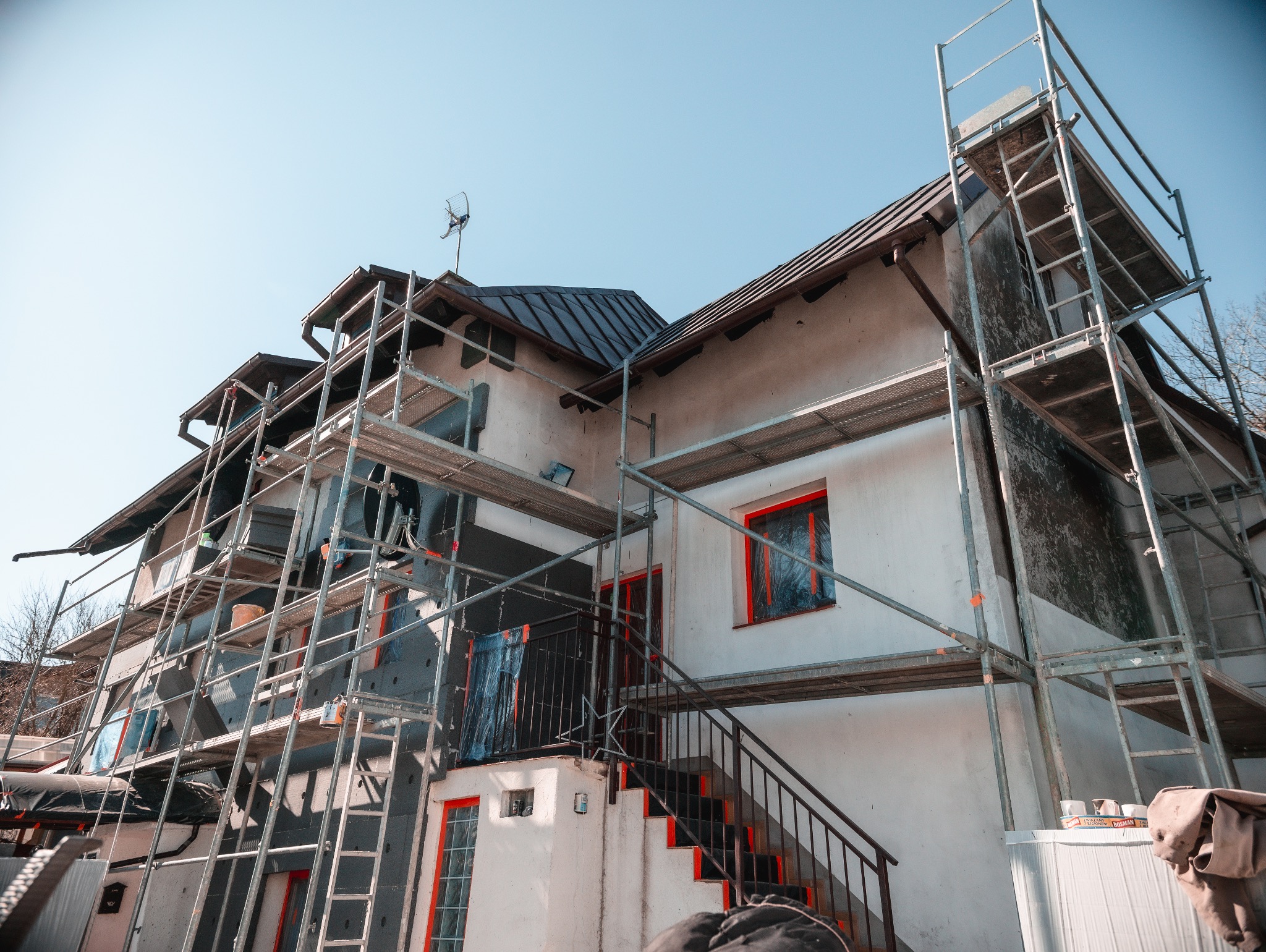 Dom w trakcie termomodernizacji, z widocznym rusztowaniem, ociepleniem ścian i zabezpieczonymi oknami. Ujęcie z dołu, przy słonecznej pogodzie, podkreśla zakres prac.