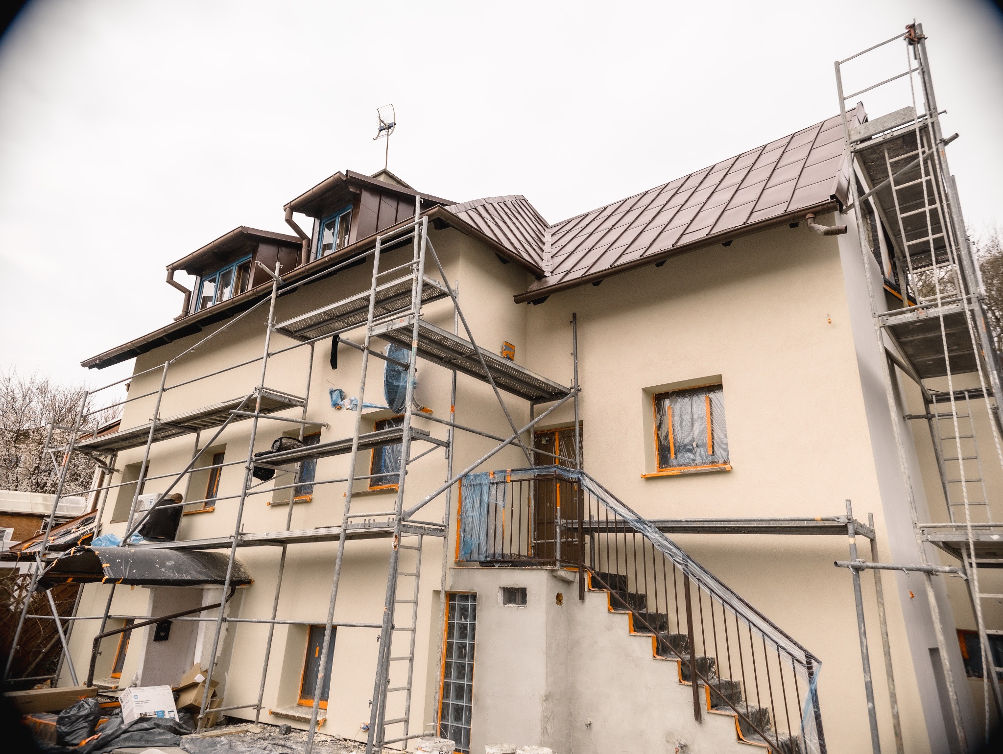 Dom w trakcie ocieplania: elewacja w jasnym odcieniu, okna zabezpieczone folią, rusztowania. Widoczny fragment dachu z brązową dachówką i schody wejściowe.