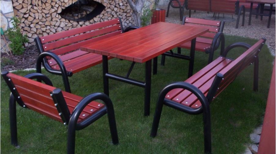 Zestaw mebli ogrodowych z drewna w odcieniach czerwieni i czarnych metalowych nóg, ustawiony na trawie, z widocznym składem drewna w tle.