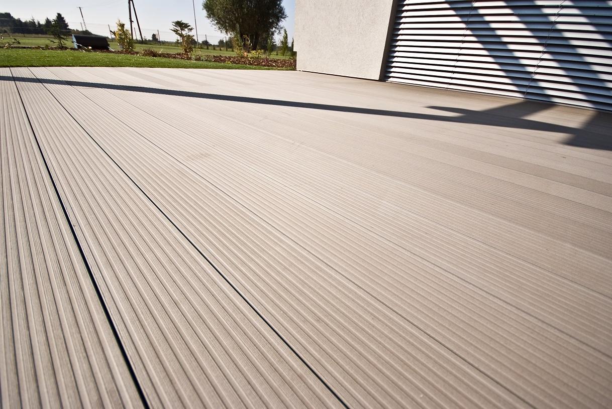 Gotowy taras z deski kompozytowej o ryflowanej powierzchni, z widocznym fragmentem budynku z żaluzjami i trawnikiem w tle.