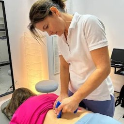 Centrum Zdrowia Rodzinnego Wocław - Kobieca Fizjoterapia - Kobieta wykonuje masaż narzędziowy dolnej części pleców pacjenta leżącego na stole w gabinecie fizjoterapii. Widoczny plakat medyczny na ścianie.