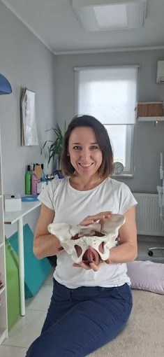 Uśmiechnięta kobieta w białej koszulce prezentuje model miednicy w jasnym gabinecie z biurkiem i oknem. Model ma anatomiczne detale.