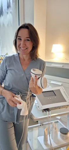 Uśmiechnięta kobieta w uniformie trzyma głowice urządzenia do zabiegów, obok stoi sprzęt kosmetyczny. W tle okno i lampka.