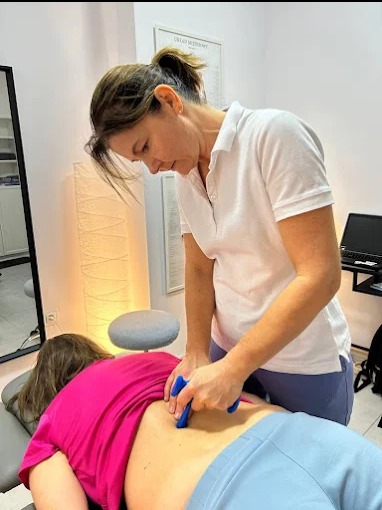 Kobieta wykonuje masaż narzędziowy dolnej części pleców pacjenta leżącego na stole w gabinecie fizjoterapii. Widoczny plakat medyczny na ścianie.