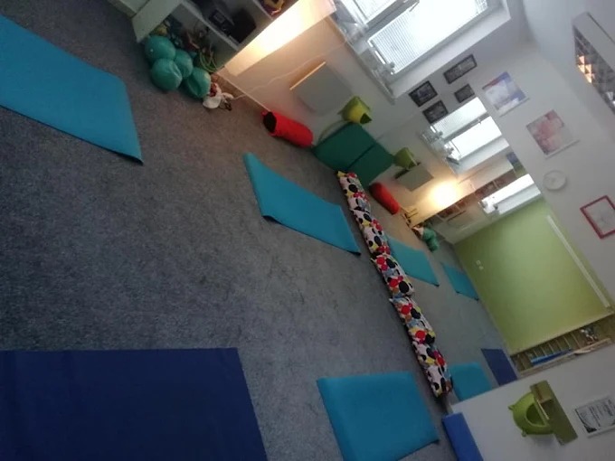 Sala do ćwiczeń z poduszkami w kształcie wałków i niebieskimi matami na szarym dywanie. Widoczne zabawki i dekoracje na ścianach, tworzące przyjazną atmosferę.