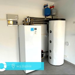 JR7 FINANZAS Robert Jędrzejczyk - Biała pompa ciepła i zbiornik buforowy w jasnym pomieszczeniu z szarą podłogą. Widoczne rury, zawory i skrzynka elektryczna na ścianie. Nowoczesna instalacja grzewcza.