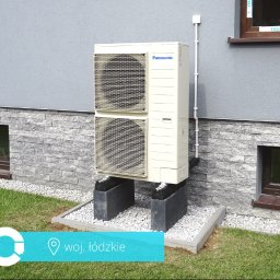 JR7 FINANZAS Robert Jędrzejczyk - Zewnętrzna jednostka klimatyzacji Panasonic, zamontowana na betonowych podstawach przy ścianie budynku z oknami i ozdobną elewacją z kamienia.