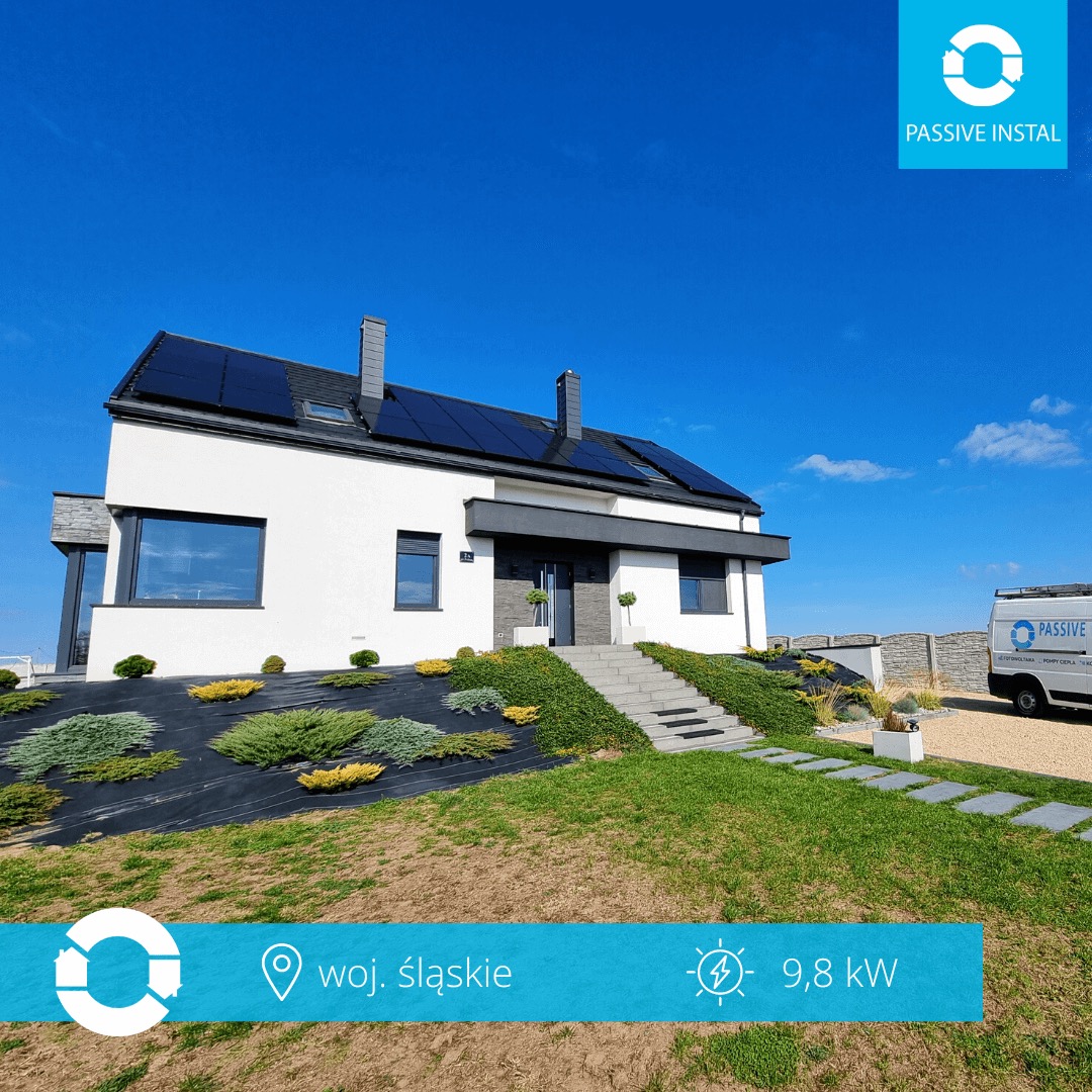 Biały dom jednorodzinny z czarnymi panelami fotowoltaicznymi na dachu, widok z dołu, błękitne niebo, zadbany ogród, bus firmy w tle.