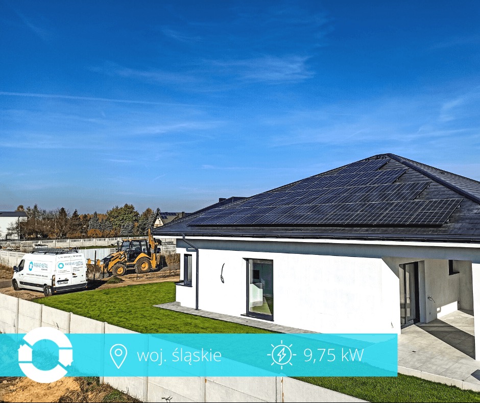 Biały dom jednorodzinny z panelami fotowoltaicznymi na dachu, na tle błękitnego nieba. Na trawniku stoi bus i koparka. Moc instalacji 9,75 kW.