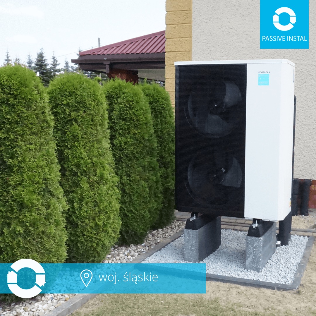 Biała pompa ciepła Hewalex na betonowych blokach, otoczona ozdobnym żwirem i żywopłotem z tuj. Widok z zewnątrz budynku, w tle fragment dachu.