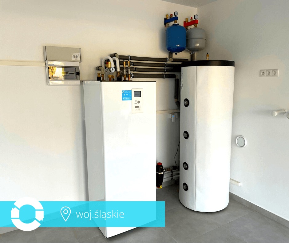 Biała pompa ciepła i zbiornik buforowy w jasnym pomieszczeniu z szarą podłogą. Widoczne rury, zawory i skrzynka elektryczna na ścianie. Nowoczesna instalacja grzewcza.