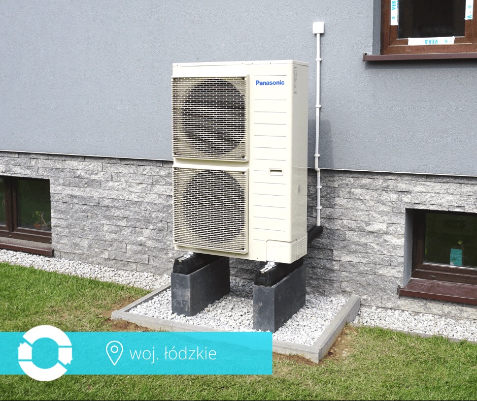 Zewnętrzna jednostka klimatyzacji Panasonic, zamontowana na betonowych podstawach przy ścianie budynku z oknami i ozdobną elewacją z kamienia.