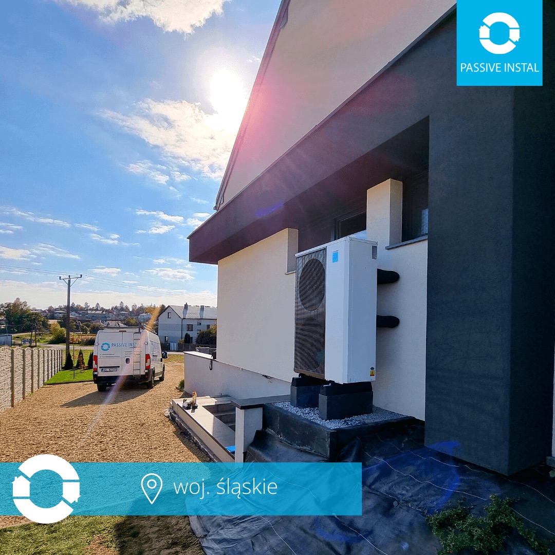 Biała pompa ciepła zamontowana na zewnątrz nowoczesnego domu z szarą elewacją, widoczna na tle błękitnego nieba i zieleni krajobrazu, obok zaparkowany bus firmy.