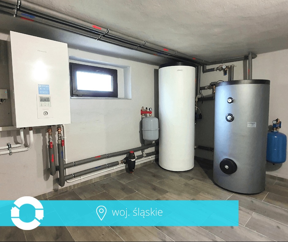 Wnętrze kotłowni z nowoczesnym piecem gazowym, zbiornikami buforowymi i instalacją rurową. Widoczne elementy automatyki i armatury. Podłoga wyłożona płytkami imitującymi drewno.