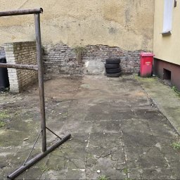 LAGARTO MAGDALENA CHORZĘPA - Zarośnięte, zaniedbane podwórko z odpadami: stary regał, opony, kosze na śmieci i czerwony pojemnik na piasek/sól. Tło: budynek z odpadającym tynkiem i ceglaną ścianą.