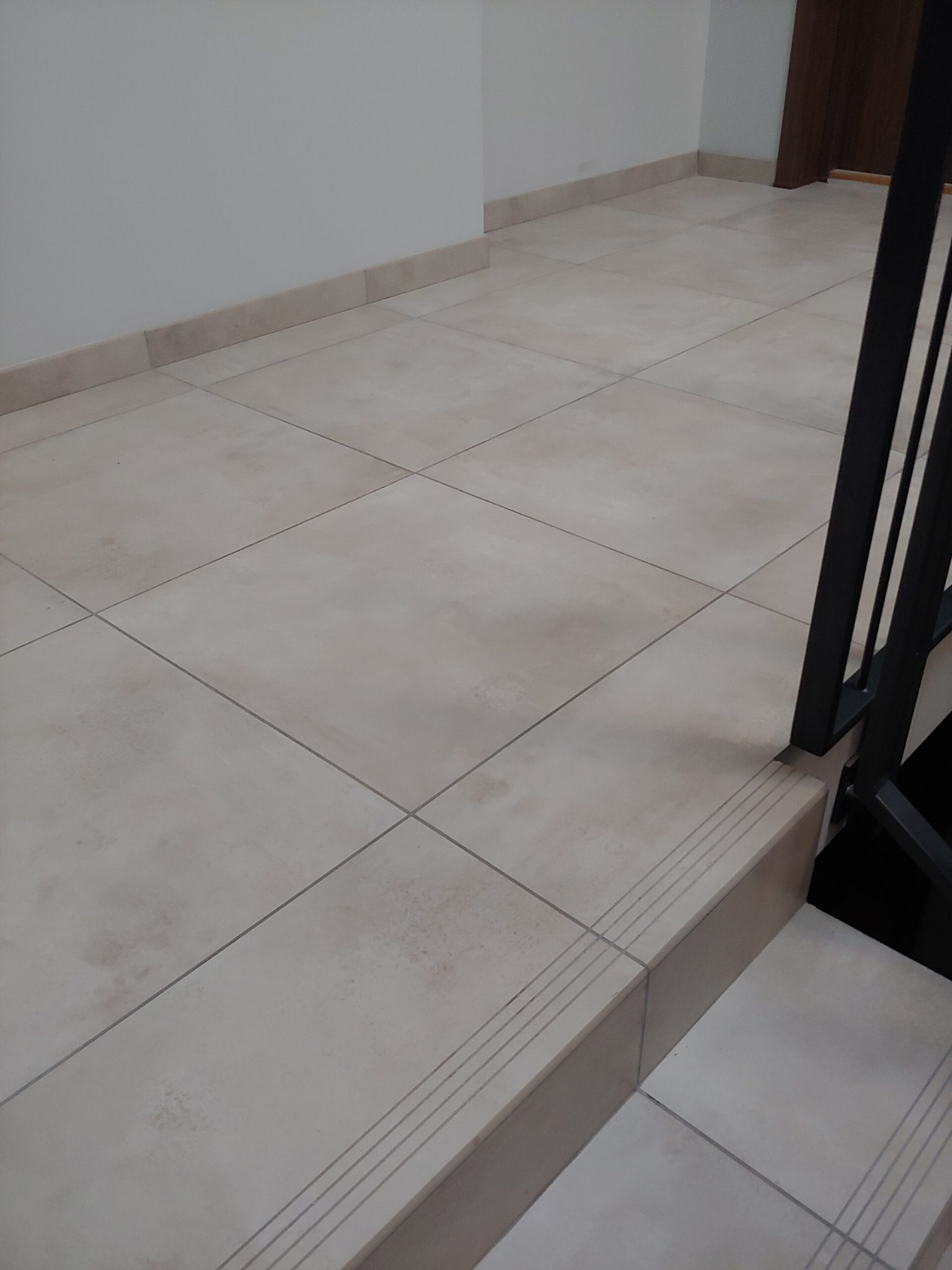 Beżowe płytki podłogowe w korytarzu, widoczne schody z czarną balustradą. Płytki ułożone równo, fuga w kolorze zbliżonym do płytek.