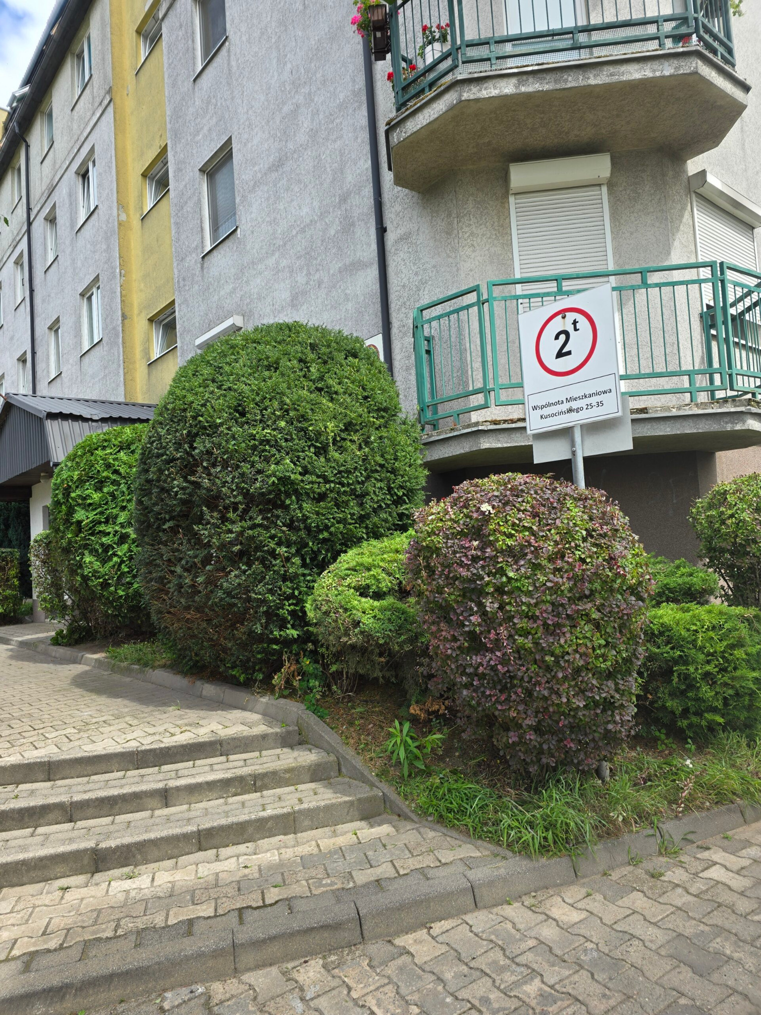 Kształtne, zielone krzewy przed budynkiem mieszkalnym, z widocznym znakiem ograniczenia tonażu. Brukowany chodnik i schody prowadzą wzdłuż roślinności.