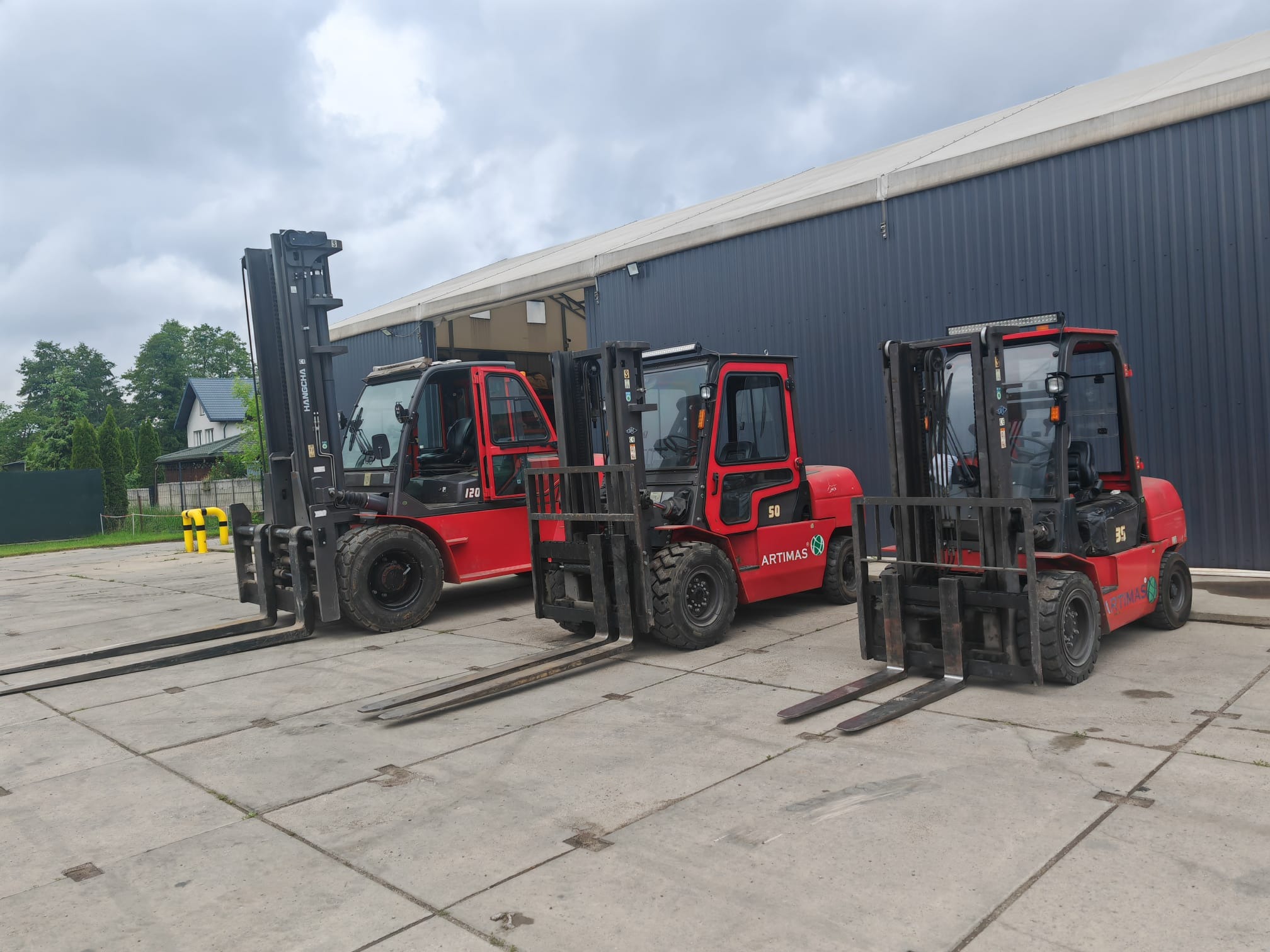 Trzy czerwone wózki widłowe marki Artimas, modele 120, 50 i 35, stoją zaparkowane na betonowym placu przed magazynem w pochmurny dzień.